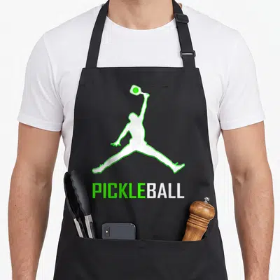 Cute player pickleball apron - White jumping person silhouette with green outline and PICKLEBALL text below — shown on a black cotton canvas apron against a white background