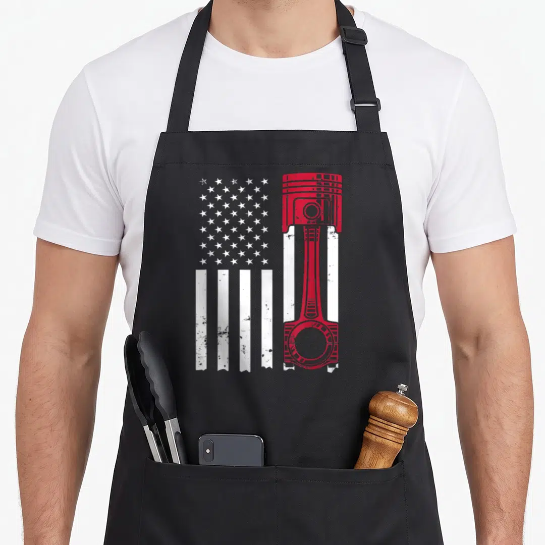 Enthusiast american flag apron - Red and white distressed USA flag with vertical engine piston replacing right side — shown on a black cotton canvas apron against a white background