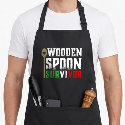 Wooden italian apron - Vertical wooden spoon with heart next to distressed WOODEN SPOON SURVIVOR text in white green — shown on a black cotton canvas apron against a white background