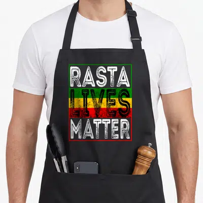 Live reggae apron - Rasta Lives Matter text in white black over green yellow red stripes inside rectangular border — shown on a black cotton canvas apron against a white background