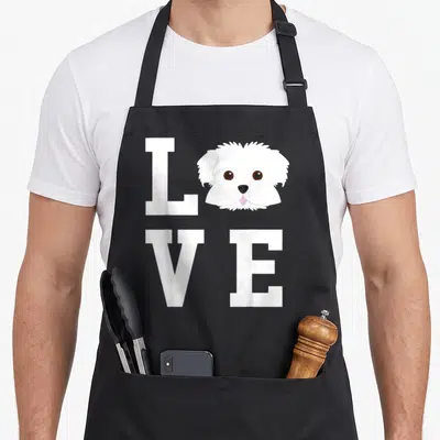Funny maltese dog apron - Cartoon white Maltese dog head replacing O in white LOVE grid with pink tongue — shown on a black cotton canvas apron against a white background