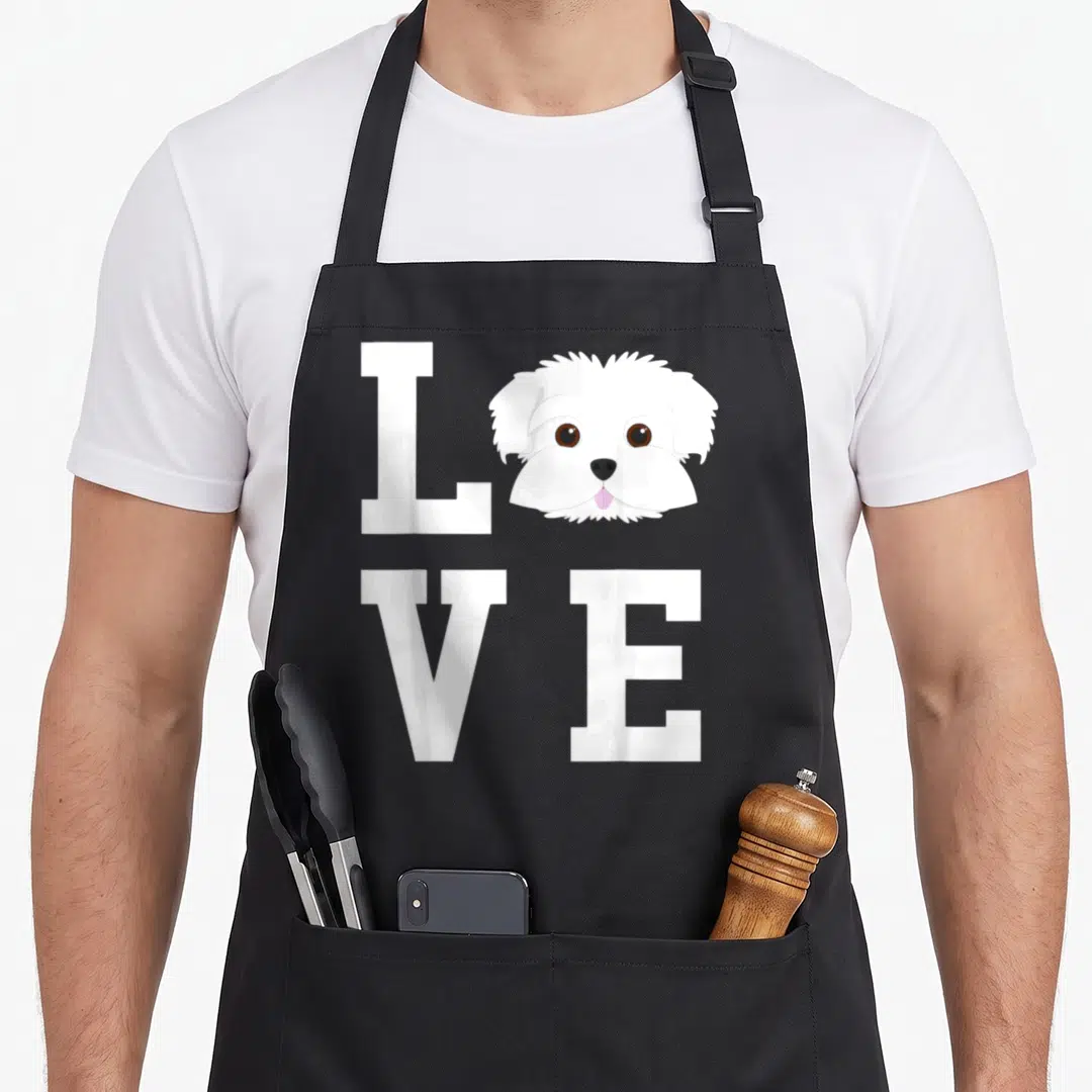 Funny maltese dog apron - Cartoon white Maltese dog head replacing O in white LOVE grid with pink tongue — shown on a black cotton canvas apron against a white background