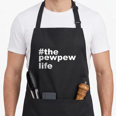 Pew husband apron - White stacked text reading #the pewpew life in bold font — shown on a black cotton canvas apron against a white background