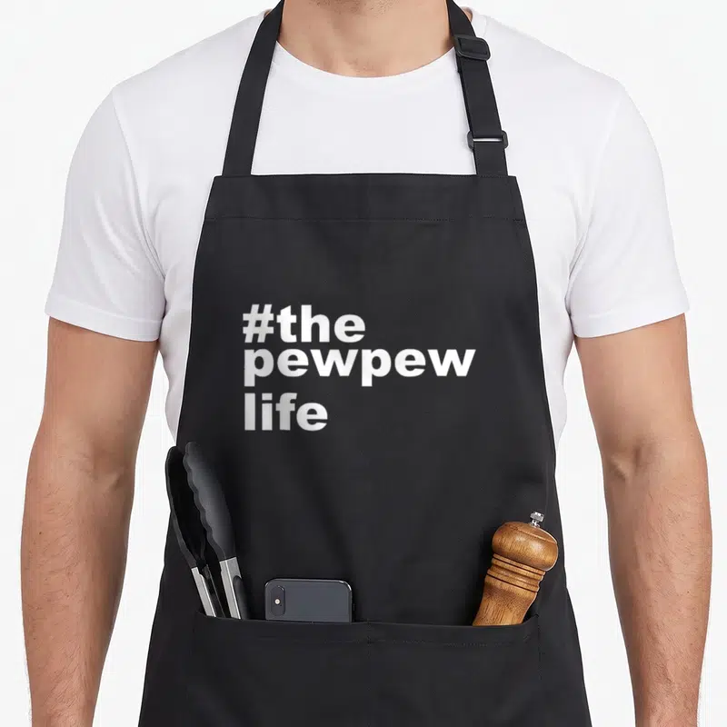 Pew husband apron - White stacked text reading #the pewpew life in bold font — shown on a black cotton canvas apron against a white background