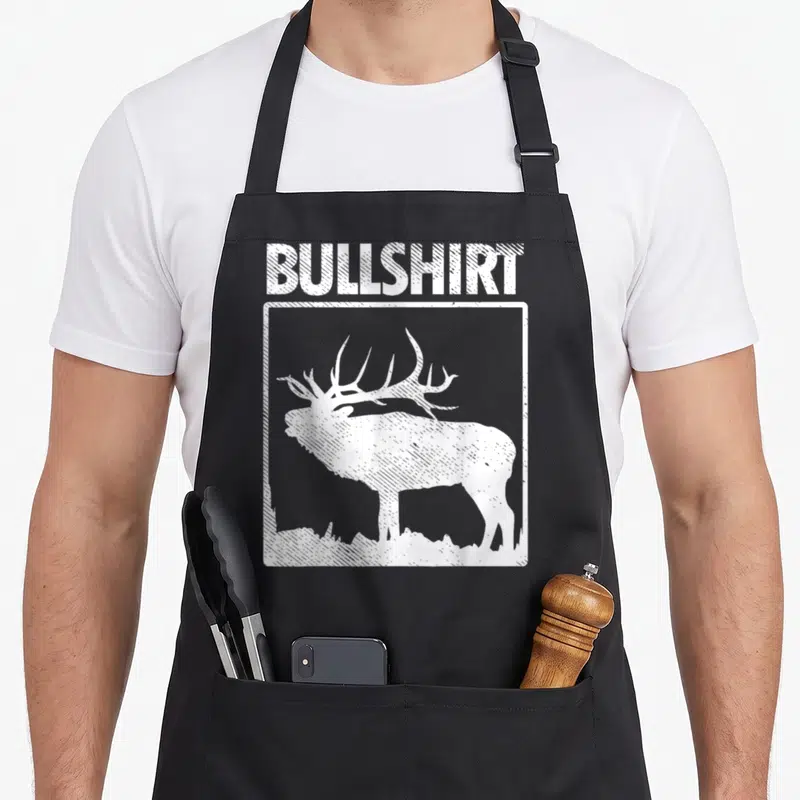 Bullshirt bow apron - Distressed white text BULLSHIRT above a square frame with an elk standing on grass — shown on a black cotton canvas apron against a white background