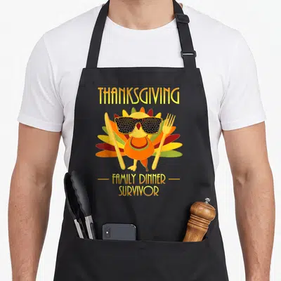 Survivor thanksgiving apron - Cartoon turkey with sunglasses flanked by knife and fork under THANKSGIVING text — shown on a black cotton canvas apron against a white background