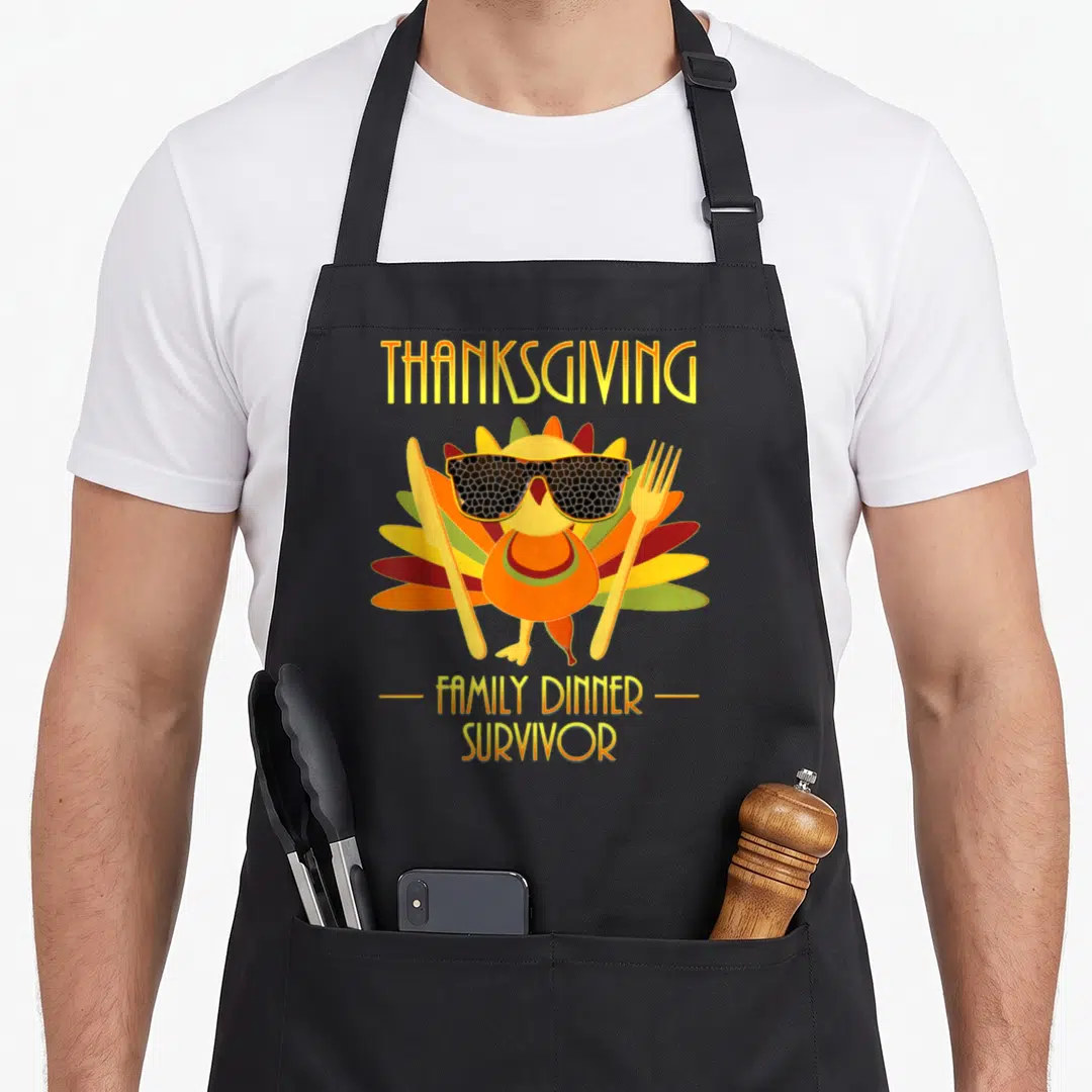 Survivor thanksgiving apron - Cartoon turkey with sunglasses flanked by knife and fork under THANKSGIVING text — shown on a black cotton canvas apron against a white background