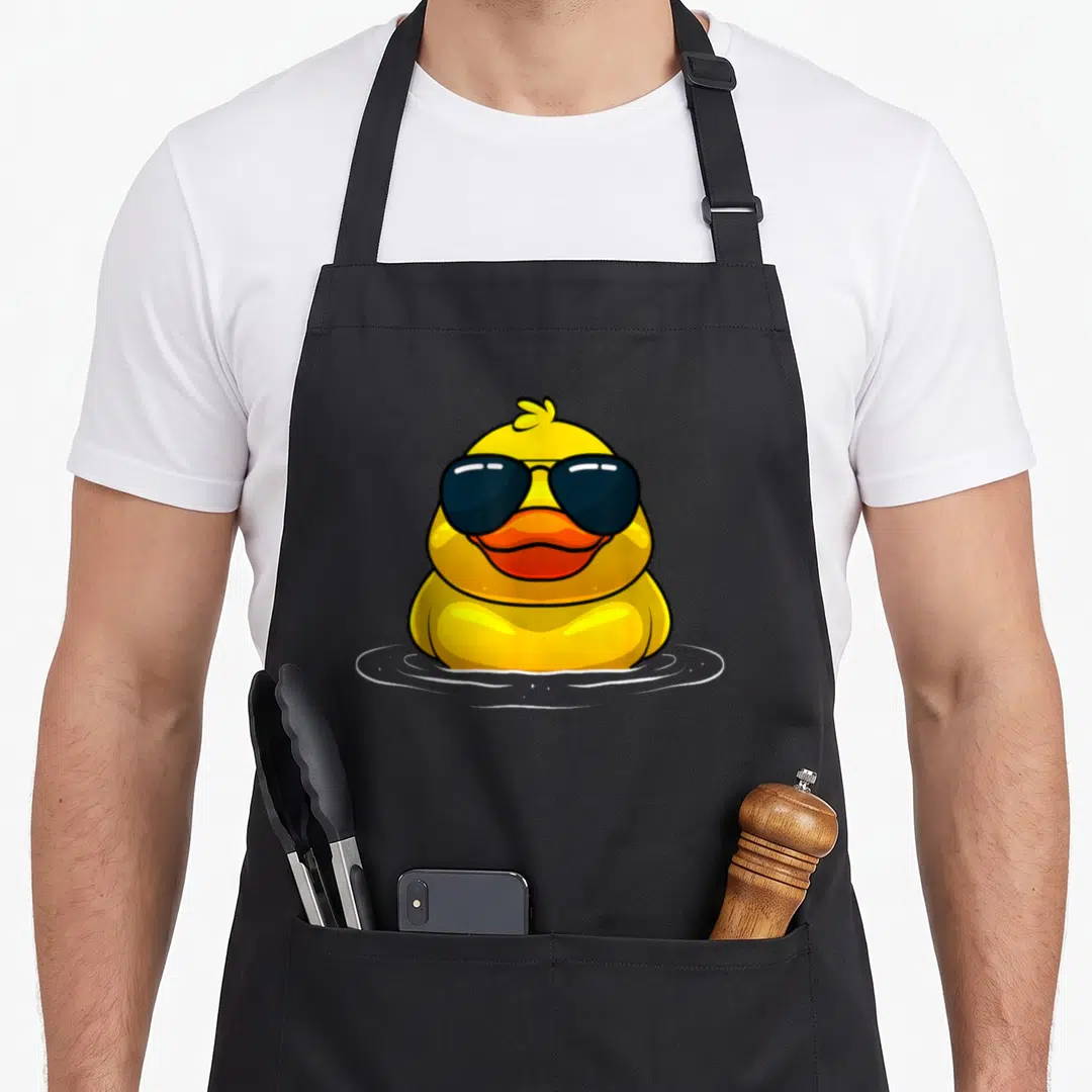 Sunglass rubber apron - Yellow cartoon duck wearing black sunglasses floating in white water ripples — shown on a black cotton canvas apron against a white background