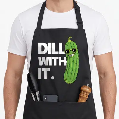 Dill joke apron - Cartoon green pickle wearing sunglasses next to white DILL WITH IT text — shown on a black cotton canvas apron against a white background