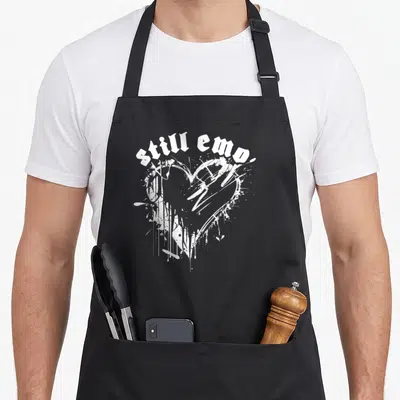Emo punk rock apron - White distressed heart made of paint splatters with arched text reading still emo — shown on a black cotton canvas apron against a white background