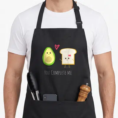 Valentine toast apron - Cartoon avocado and toast standing with pink heart and YOU COMPLETE ME text — shown on a black cotton canvas apron against a white background