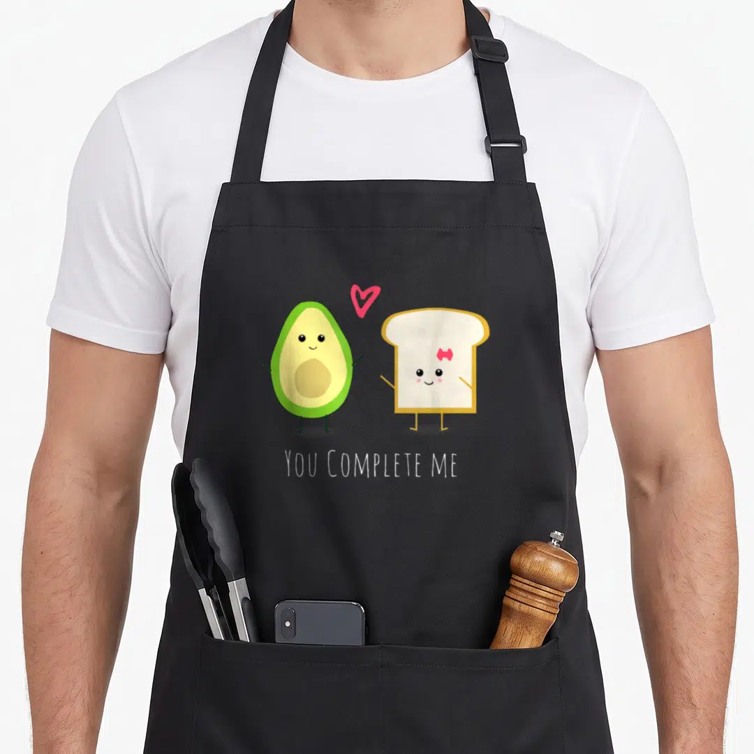 Valentine toast apron - Cartoon avocado and toast standing with pink heart and YOU COMPLETE ME text — shown on a black cotton canvas apron against a white background