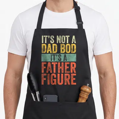 Father figure dad bod apron - Stacked retro text IT'S NOT A DAD BOD IT'S A FATHER FIGURE in beige yellow and orange-red — shown on a black cotton canvas apron against a white background