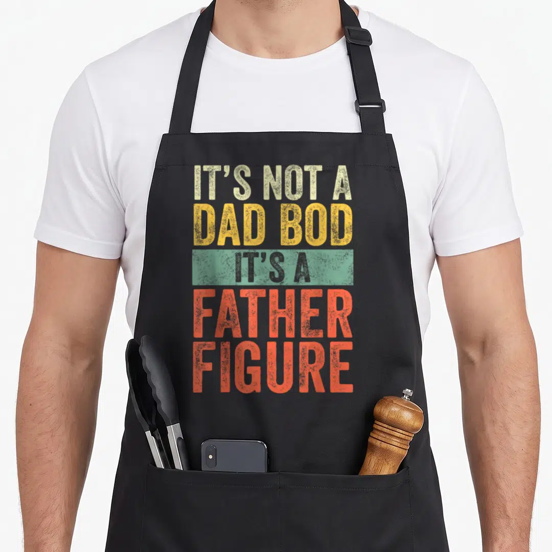 Father figure dad bod apron - Stacked retro text IT'S NOT A DAD BOD IT'S A FATHER FIGURE in beige yellow and orange-red — shown on a black cotton canvas apron against a white background