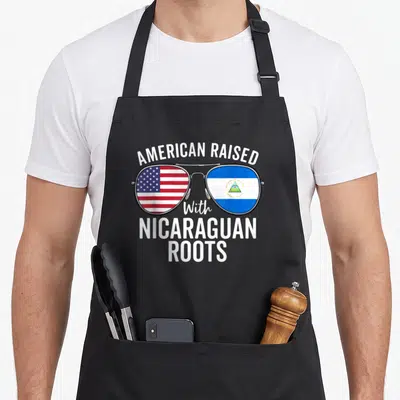 Raised american flag apron - Sunglasses with USA and Nicaragua flags in lenses under AMERICAN RAISED With NICARAGUAN — shown on a black cotton canvas apron against a white background
