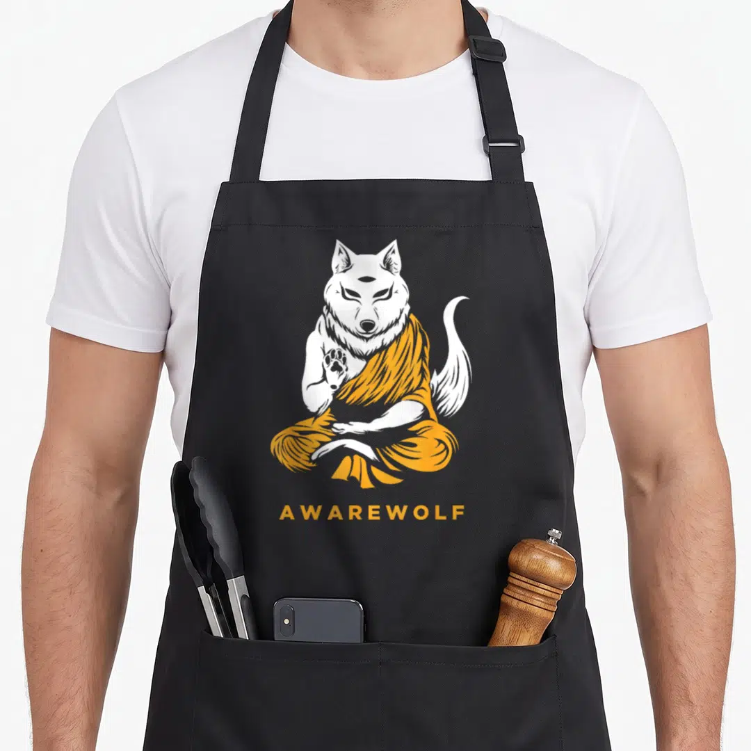 Yoga werewolf apron - White wolf in orange monk robe meditating above orange AWAREWOLF text — shown on a black cotton canvas apron against a white background