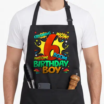 6th comic birthday apron - Red number 6 surrounded by comic text CRUSHING WOW BOOM AWESOME BIRTHDAY BOY with stars — shown on a black cotton canvas apron against a white background