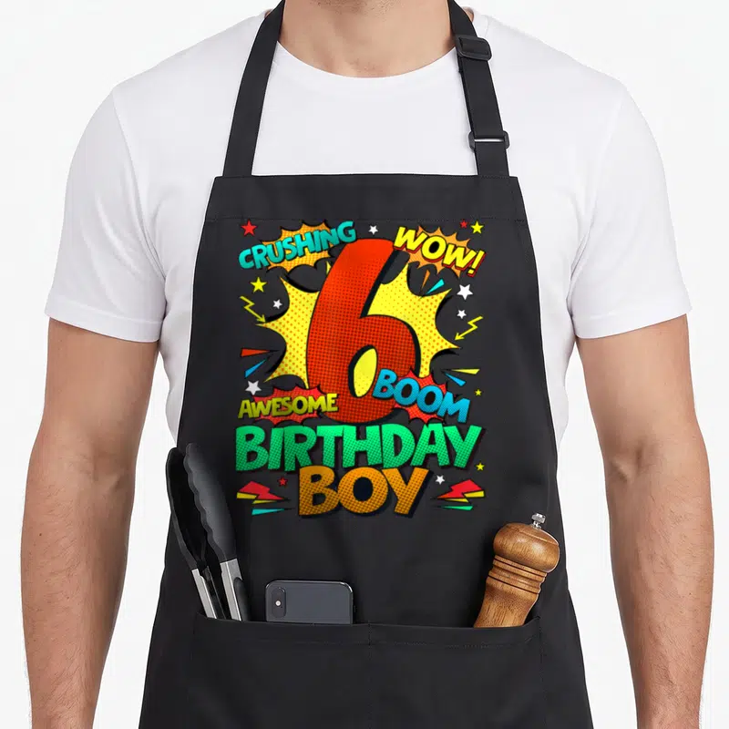 6th comic birthday apron - Red number 6 surrounded by comic text CRUSHING WOW BOOM AWESOME BIRTHDAY BOY with stars — shown on a black cotton canvas apron against a white background