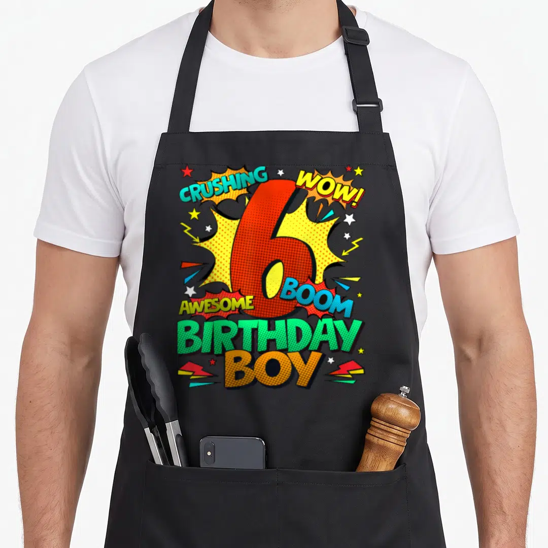 6th comic birthday apron - Red number 6 surrounded by comic text CRUSHING WOW BOOM AWESOME BIRTHDAY BOY with stars — shown on a black cotton canvas apron against a white background