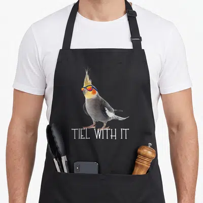 Teil bird apron - Cockatiel wearing blue sunglasses above white TIEL WITH IT text — shown on a black cotton canvas apron against a white background