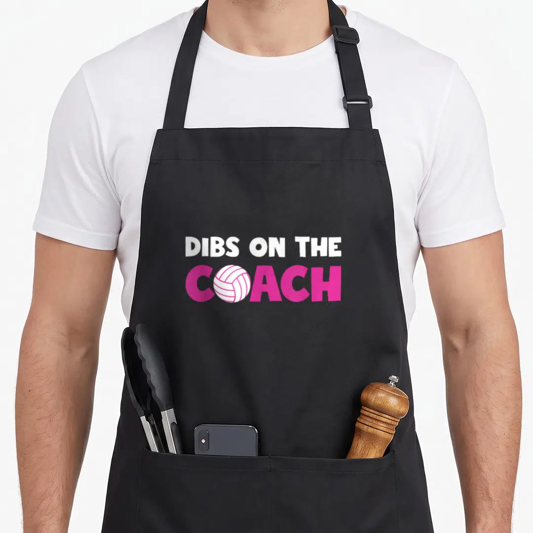 Volleyball wife apron - DIBS ON THE COACH text with pink letters and white volleyball replacing the O — shown on a black cotton canvas apron against a white background