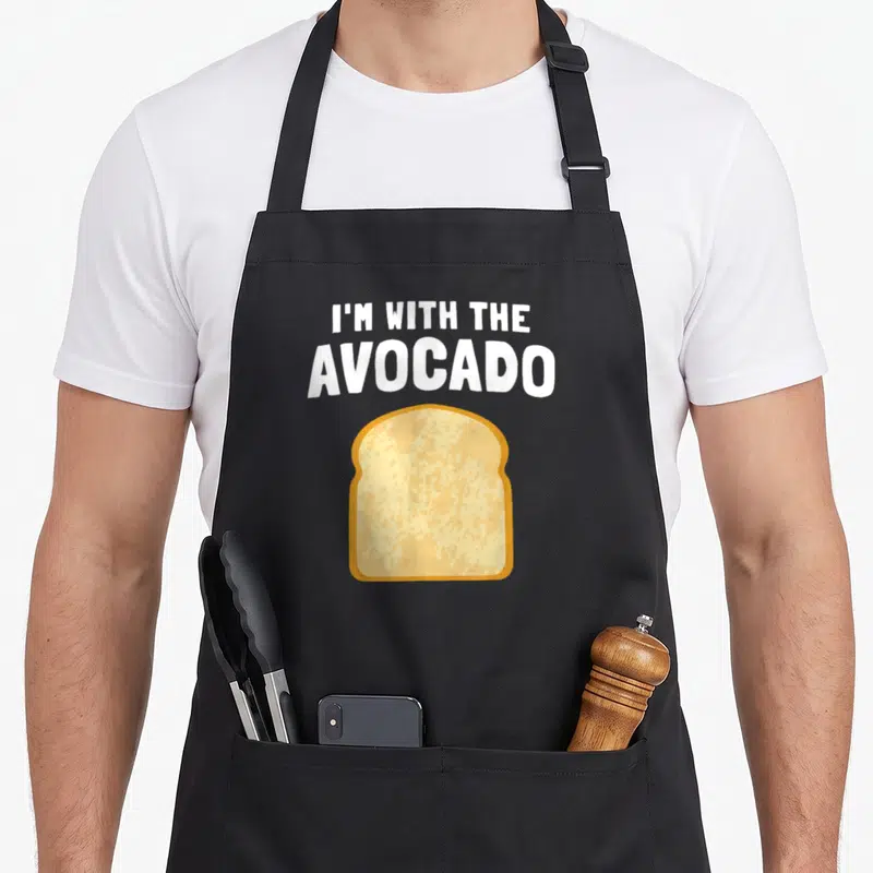 Avocado toast apron - I'M WITH THE AVOCADO text above cartoon toast slice with two pockets — shown on a black cotton canvas apron against a white background