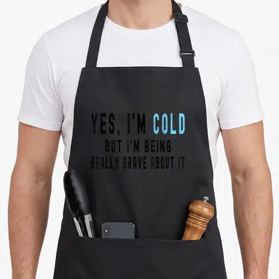 Cold novelty apron - Stacked text reading Yes I'm Cold But I'm Being Really Brave About It in black and blue distressed — shown on a black cotton canvas apron against a white background