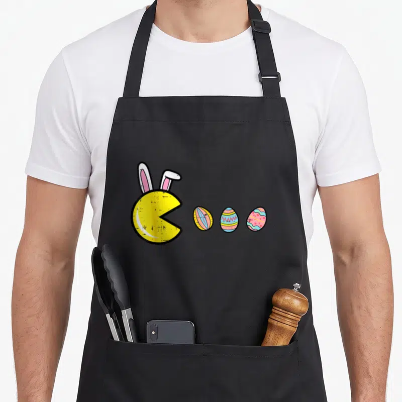 Eating easter egg apron - Yellow Pac-Man character with pink bunny ears facing three blue and pink decorated Easter eggs — shown on a black cotton canvas apron against a white background