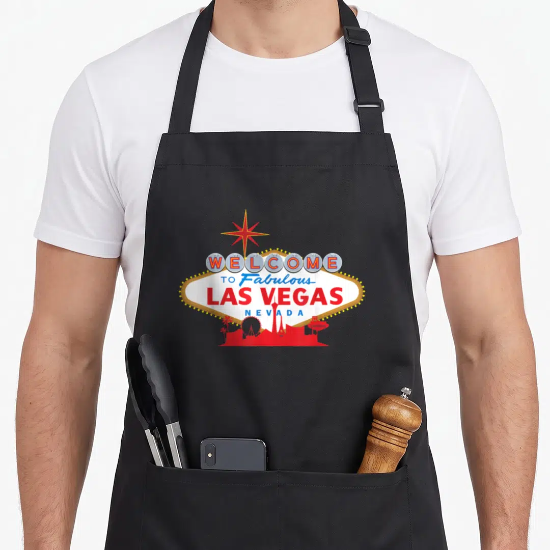 Las holiday apron - Retro Las Vegas sign with star, red skyline silhouette, and welcome text in red white blue — shown on a black cotton canvas apron against a white background