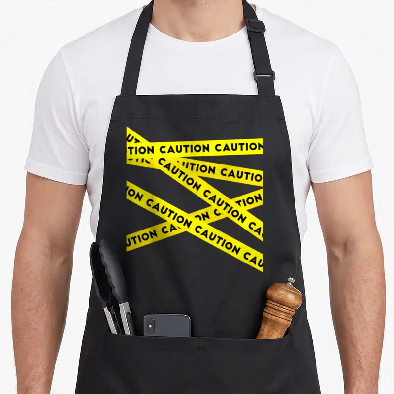 Caution modern apron - Yellow strips with black CAUTION text overlapping on a dark background — shown on a black cotton canvas apron against a white background