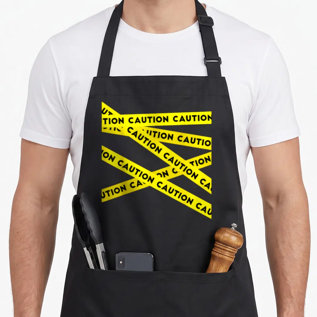 Caution modern apron - Yellow strips with black CAUTION text overlapping on a dark background — shown on a black cotton canvas apron against a white background