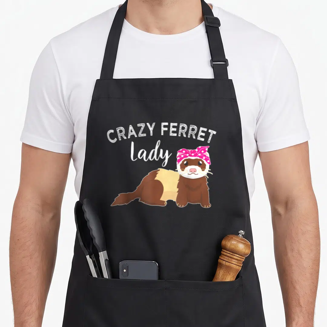 Funny ferret animal apron - Cartoon brown ferret wearing a pink polka dot bandana below white text reading CRAZY FERRET — shown on a black cotton canvas apron against a white background