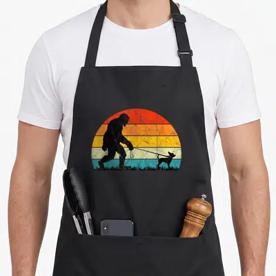 Walking dog training apron - Bigfoot silhouette walking small dog on leash against striped retro sunset — shown on a black cotton canvas apron against a white background