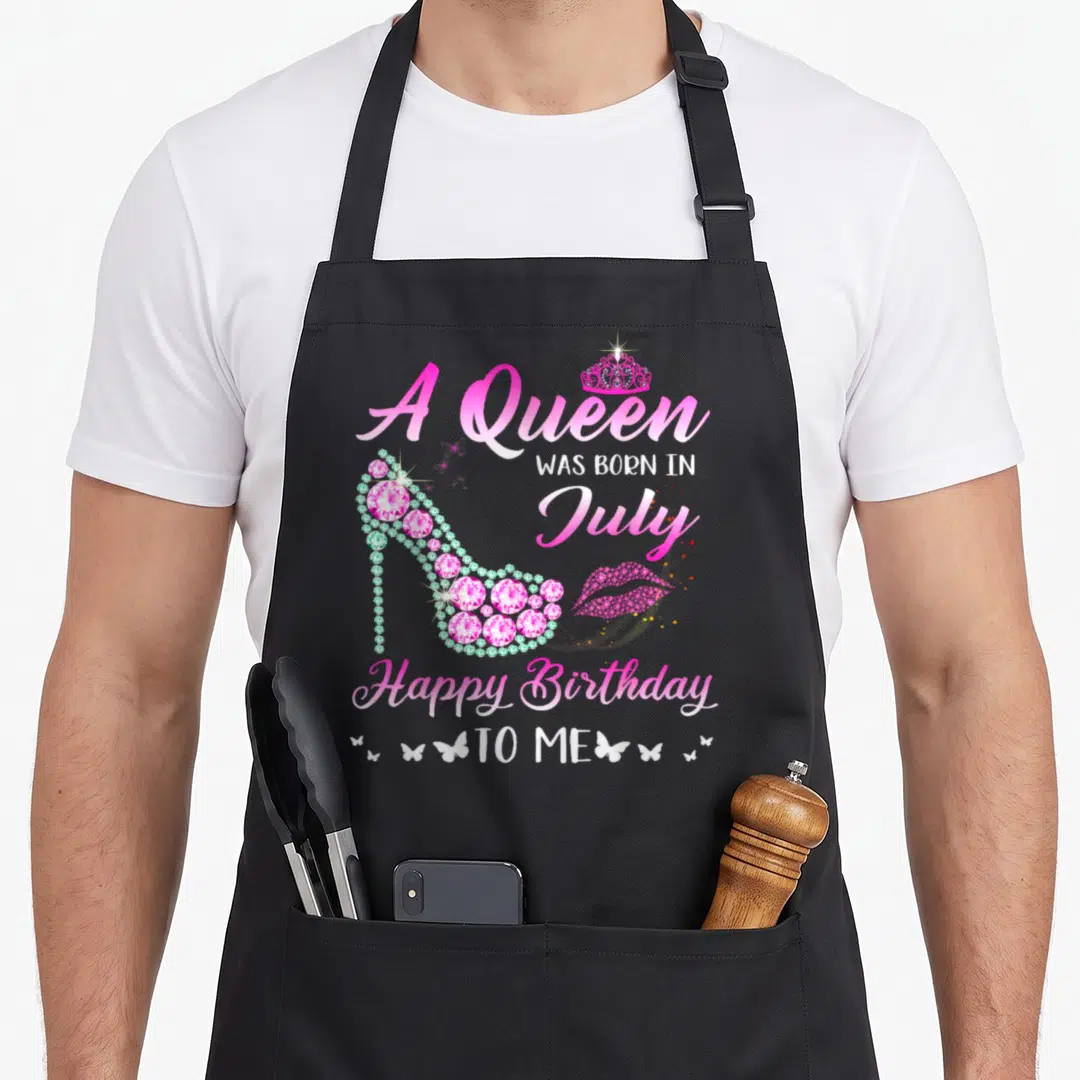 July birthday baking queen apron - Pink high heel shoe, crown, lips, and July birthday text on white background — shown on a black cotton canvas apron against a white background