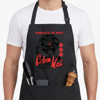 Train mercy apron - Red Cobra Kai script, black cobra, kanji, three fist icons, and Victory Blvd address — shown on a black cotton canvas apron against a white background