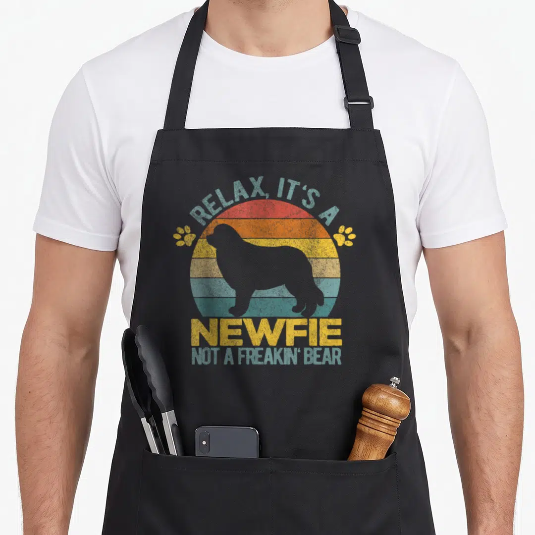 Newfie owner dog apron - Retro sun graphic with grey dog silhouette and text Relax Its a Newfie Not a Freakin Bear — shown on a black cotton canvas apron against a white background