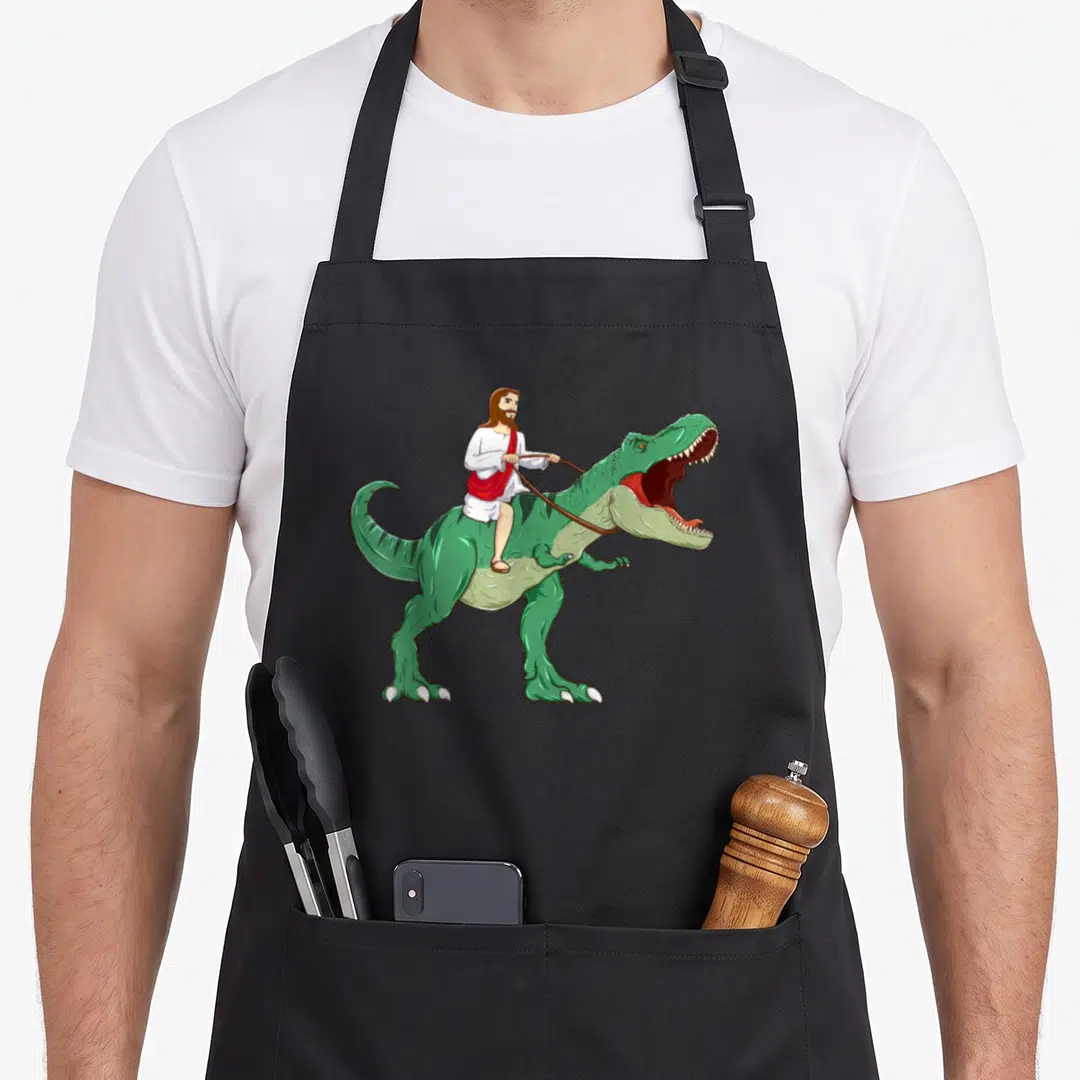 Parody meme apron - Cartoon Jesus in white and red robes riding a green T-Rex holding brown reins — shown on a black cotton canvas apron against a white background
