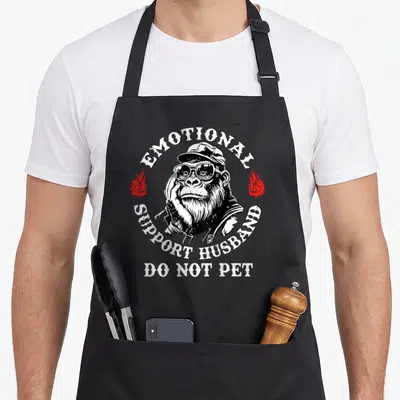 Monkey husband apron - Chimpanzee in glasses and jacket with emotional support husband text and red flames — shown on a black cotton canvas apron against a white background