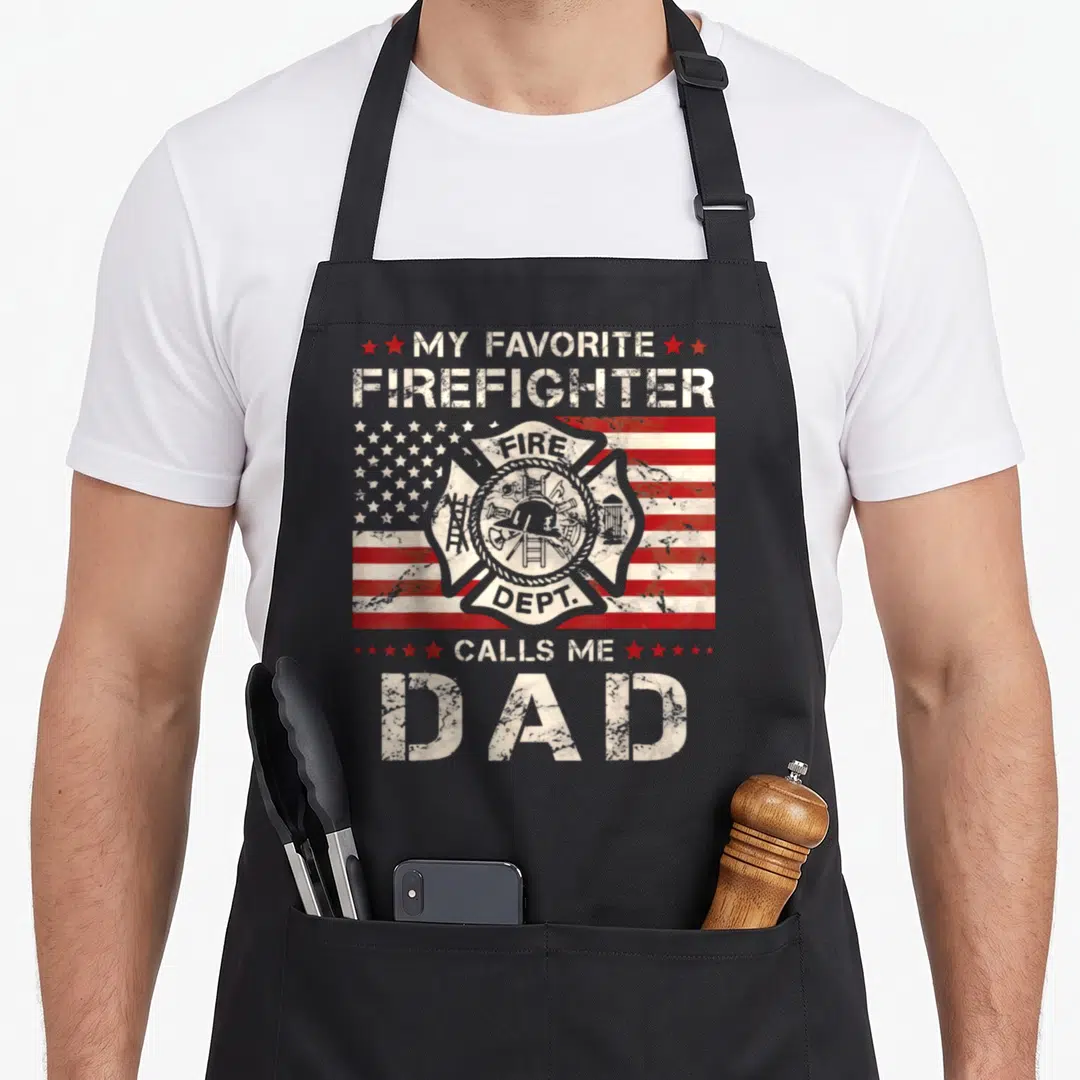 Call firefighter aprons - Distressed text My Favorite Firefighter Calls Me Dad over Maltese cross and US flag — shown on a black cotton canvas apron against a white background