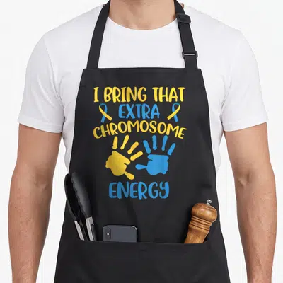 Chromosome an extra apron - I Bring That Extra Chromosome Energy text with yellow blue handprints and ribbons — shown on a black cotton canvas apron against a white background