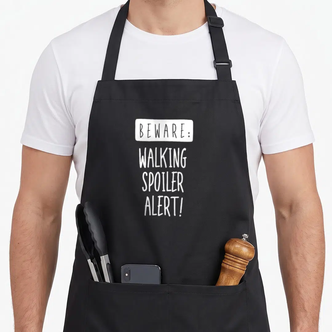 Beware walking apron - White stacked text reading Beware Walking Spoiler Alert on black background — shown on a black cotton canvas apron against a white background