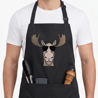 Shade moose apron - Cartoon moose head in beige and brown wearing black sunglasses — shown on a black cotton canvas apron against a white background