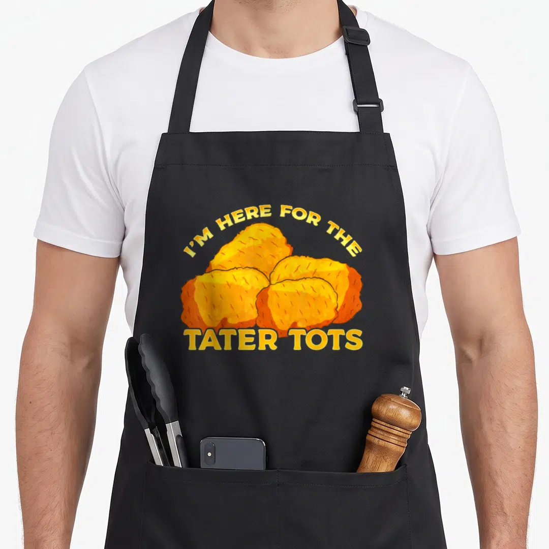 Tot funny tater apron - Five yellow and orange cartoon tater tots with arched text I'M HERE FOR THE TATER TOTS — shown on a black cotton canvas apron against a white background