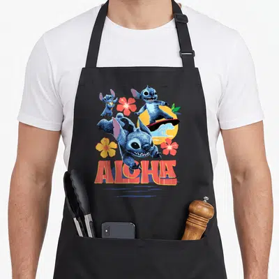 Live tropical print aprons - Three blue characters near yellow sun and wave with red ALOHA text and hibiscus flowers — shown on a black cotton canvas apron against a white background