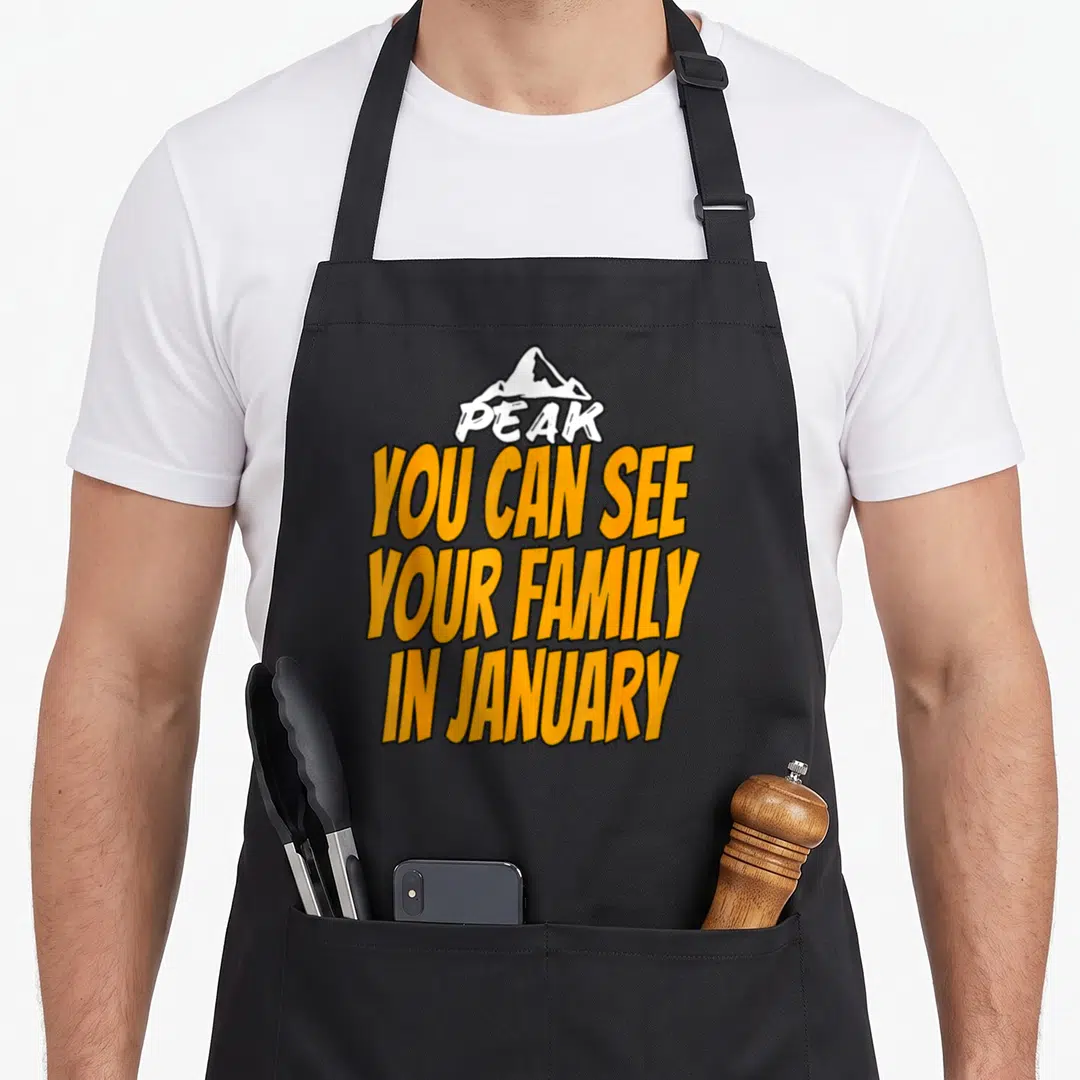 Welcome family aprons - White mountain peak graphic above orange text reading PEAK YOU CAN SEE YOUR FAMILY IN JANUARY — shown on a black cotton canvas apron against a white background