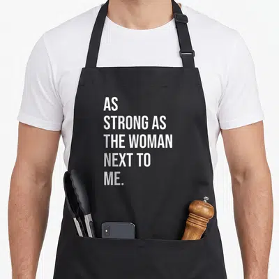 Feminism pro apron - White stacked text reading As Strong As The Woman Next To Me on black fabric — shown on a black cotton canvas apron against a white background