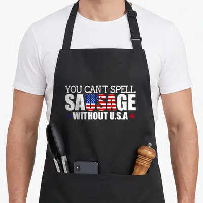 Patriotism sausage apron - Distressed text reading YOU CAN'T SPELL SAUSAGE WITHOUT U.S.A with flag pattern in middle — shown on a black cotton canvas apron against a white background