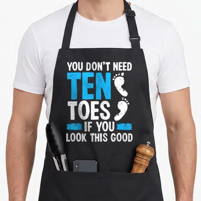 Toe survivor apron - Stacked text YOU DON'T NEED TEN TOES with two white footprints and blue brush strokes — shown on a black cotton canvas apron against a white background