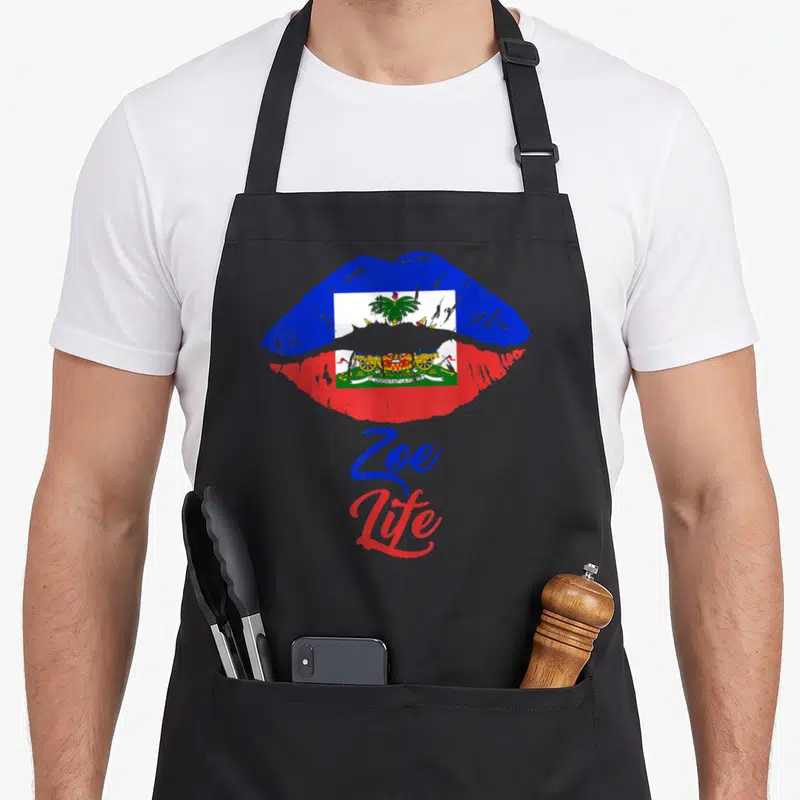 May pride apron - Cartoon lips filled with Haitian flag and Zoe Life text in blue and red script — shown on a black cotton canvas apron against a white background