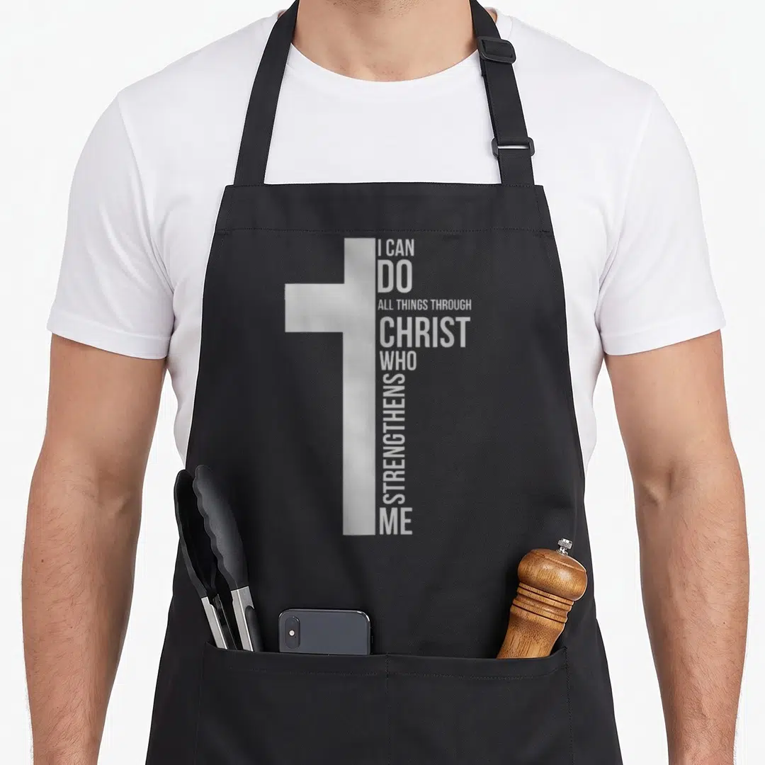 Through cross apron - White cross graphic with stacked typography reading I CAN DO ALL THINGS THROUGH CHRIST WHO — shown on a black cotton canvas apron against a white background